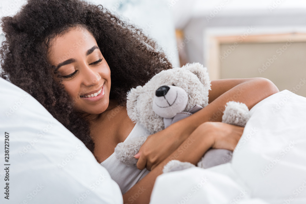 close up view of smiling african american curly girl laying in bed with teddy bear at home