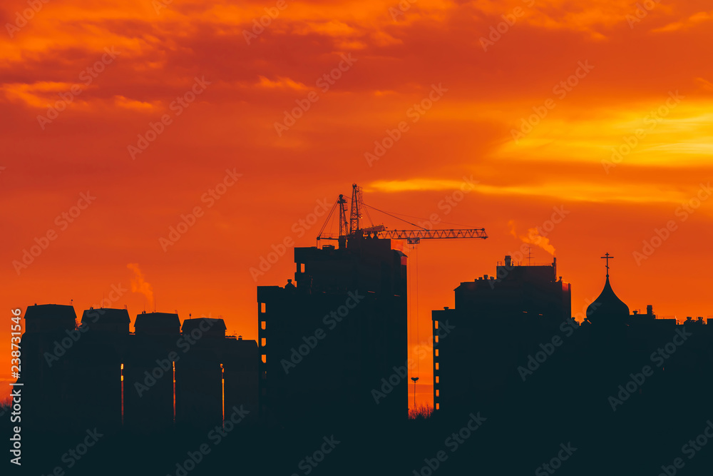 Cityscape with vivid fiery dawn. Amazing warm dramatic cloudy sky above dark silhouettes of city buildings. Orange sunlight. Atmospheric background of sunrise in overcast weather. Copy space.