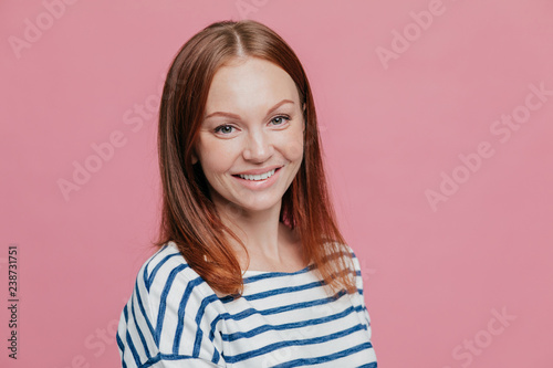 Sideways shot of pleasant looking happy brown haired woman with straight hair, healthy skin, pleasant smile, dressed in casual striped sweater, isolated over pink background. Emotions concept
