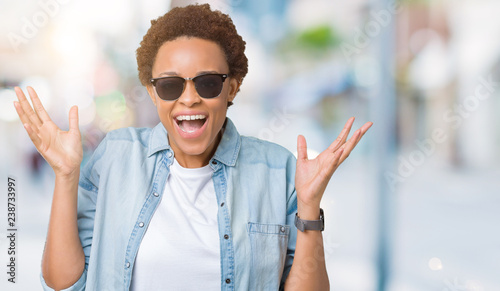 Beautiful young african american woman wearing sunglasses over isolated background celebrating mad and crazy for success with arms raised and closed eyes screaming excited. Winner concept