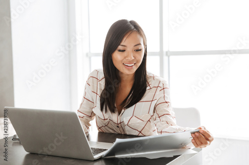 Image of young asian woman 20s working on laptop, while sitting at table in flat
