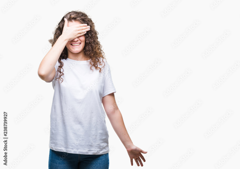 Beautiful brunette curly hair young girl wearing casual t-shirt over isolated background smiling and laughing with hand on face covering eyes for surprise. Blind concept.