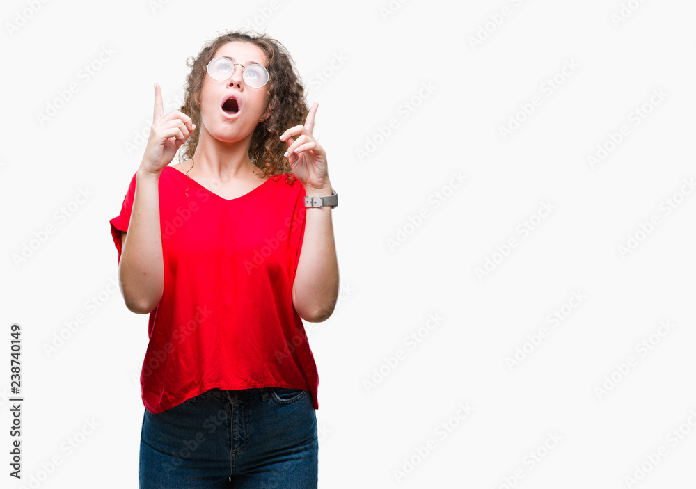 Beautiful brunette curly hair young girl wearing glasses over isolated background amazed and surprised looking up and pointing with fingers and raised arms. Beautiful brunette curly hair young girl wearing glasses over isolated background amazed and surprised looking up and pointing with fingers and raised arms.