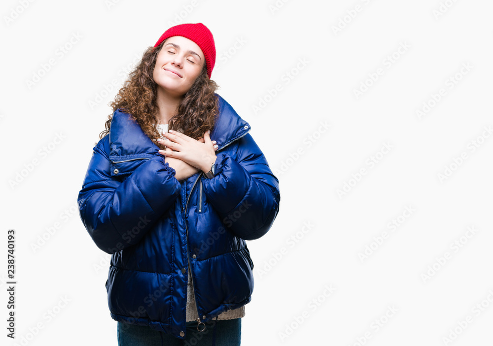 Beautiful young brunette curly hair girl wearing winter coat, wool cap and sweater over isolated background smiling with hands on chest with closed eyes and grateful gesture on face. Health concept. Beautiful young brunette curly hair girl wearing winter coat, wool cap and sweater over isolated background smiling with hands on chest with closed eyes and grateful gesture on face. Health concept.