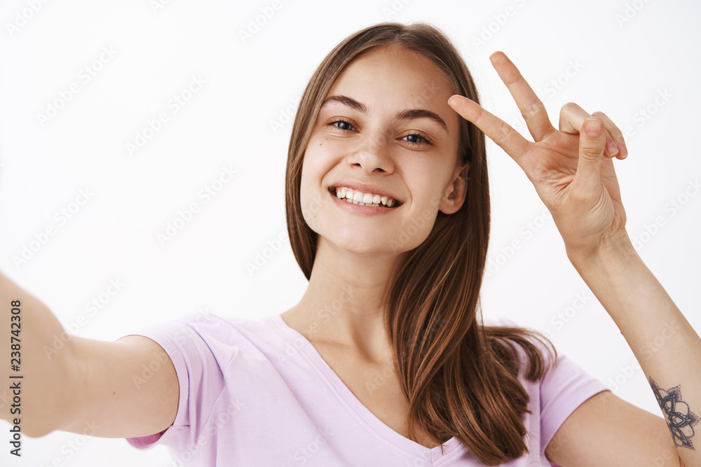 Saying hi to internet followers. Charming friendly-looking joyful girl with brown hair smiling broadly showing peace or victory sign near face while taking selfie with smartphone over grey wall Saying hi to internet followers. Charming friendly-looking joyful girl with brown hair smiling broadly showing peace or victory sign near face while taking selfie with smartphone over grey wall