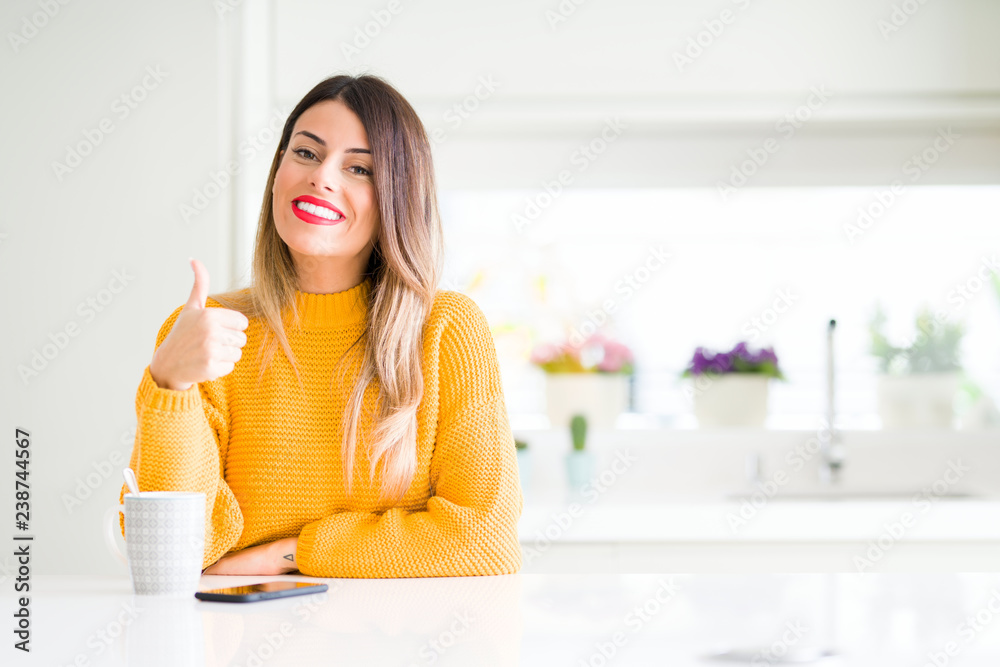 Young beautiful woman drinking a cup of coffee at home doing happy thumbs up gesture with hand. Approving expression looking at the camera showing success.