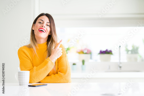 Young beautiful woman drinking a cup of coffee at home cheerful with a smile of face pointing with hand and finger up to the side with happy and natural expression on face