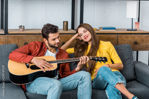 young handsome man sitting on sofa in living room and playing guitar for attractive girlfriend