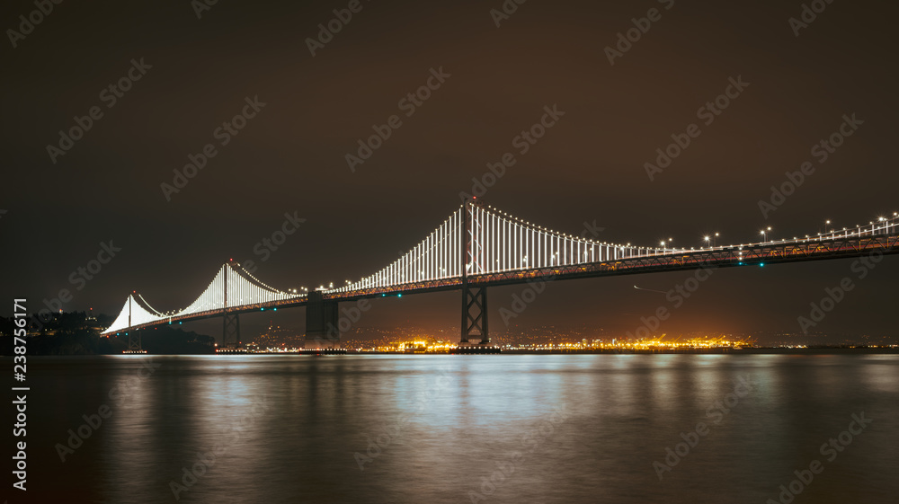 The Bay Bridge lit up at night with reflections on the Bay