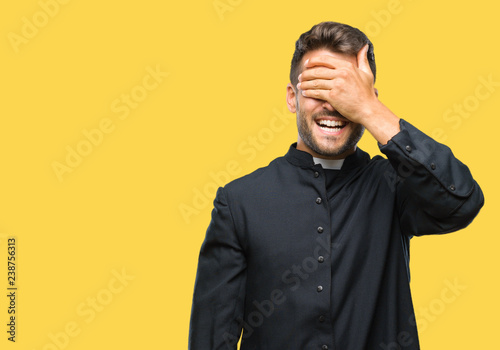 Young catholic christian priest man over isolated background smiling and laughing with hand on face covering eyes for surprise. Blind concept.