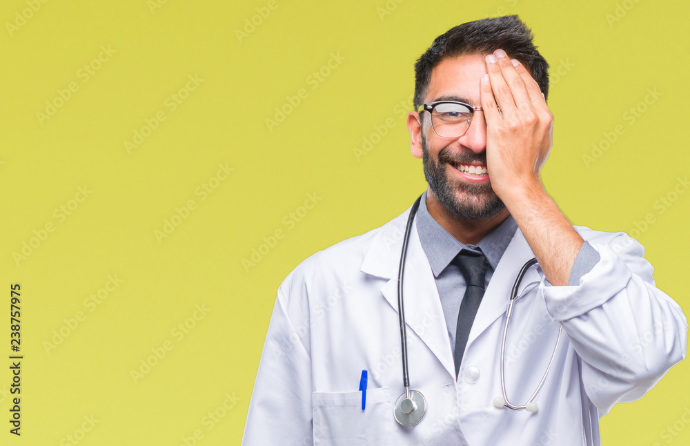 Adult hispanic doctor man over isolated background covering one eye with hand with confident smile on face and surprise emotion. Adult hispanic doctor man over isolated background covering one eye with hand with confident smile on face and surprise emotion.