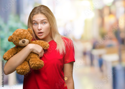 Young caucasian woman holding cute teddy bear over isolated background scared in shock with a surprise face  afraid and excited with fear expression