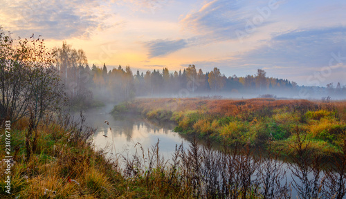 Foggy autumn landscape with small forest river.Autumnal hazy early morning.Twilight.Calm stream of river flowing between the woods and meadows covered by fog.Colorful clouds in sky at sunrise.