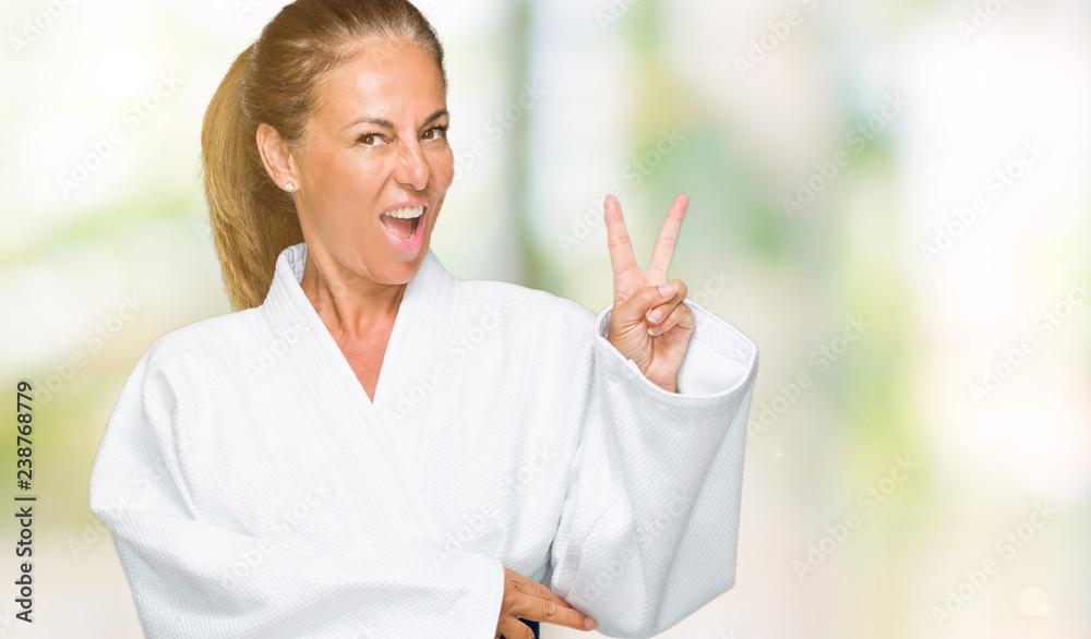 Fototapeta premium Middle age adult woman wearing karate kimono uniform over isolated background smiling with happy face winking at the camera doing victory sign. Number two.
