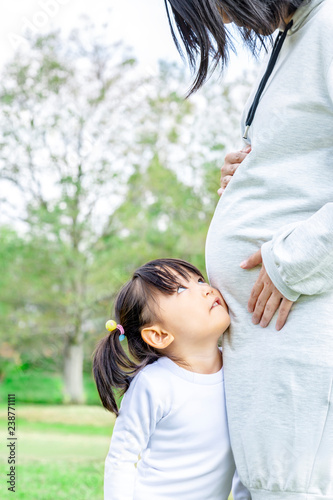 公園で妊娠しているお母さんの大きなお腹に顔を当て見上げる幼い女の子。親子、家族、愛、幸せ、妊娠イメージ