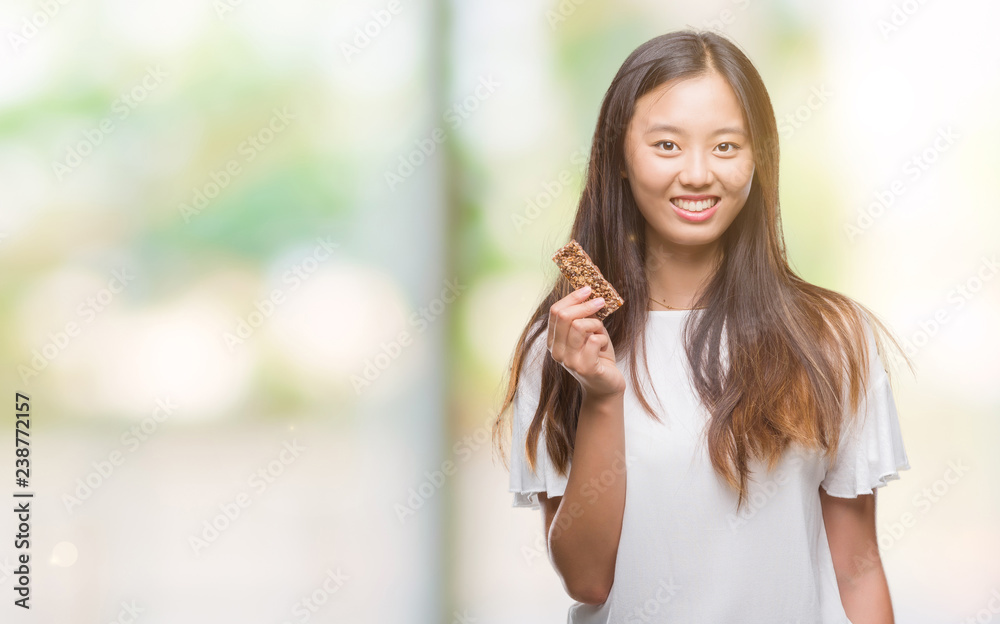 Young asian woman eating chocolate energetic bar over isolated background with a happy face standing and smiling with a confident smile showing teeth