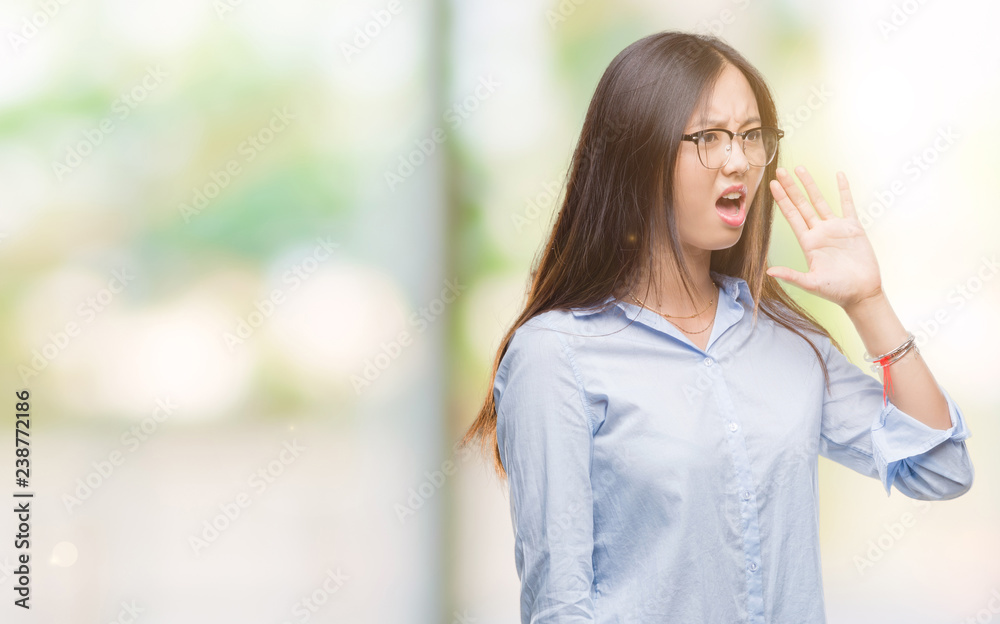 Young asian business woman wearing glasses over isolated background shouting and screaming loud to side with hand on mouth. Communication concept. Young asian business woman wearing glasses over isolated background shouting and screaming loud to side with hand on mouth. Communication concept.