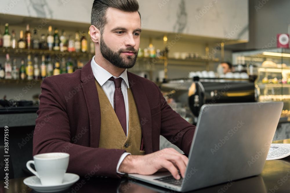 handsome businessman sitting at table with cup of coffee and typing on laptop in restaurant handsome businessman sitting at table with cup of coffee and typing on laptop in restaurant