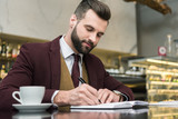 handsome businessman in formal wear sitting and writing in notebook at table in restaurant