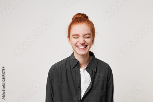 Cheerful resilient red-haired girl with a bun at her head laughs sincerely, having closed her eyes, dressed in a simple black shirt, is isolated on white background