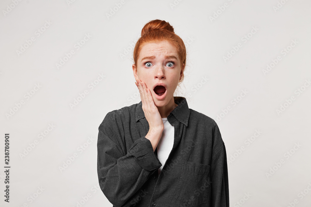 Red-haired woman looks shocked frightened of the catastrophe, crash, disaster, effects of her actions. Scared girl stands with her eyes and mouth wide open holding her palm near her cheek. Red-haired woman looks shocked frightened of the catastrophe, crash, disaster, effects of her actions. Scared girl stands with her eyes and mouth wide open holding her palm near her cheek.