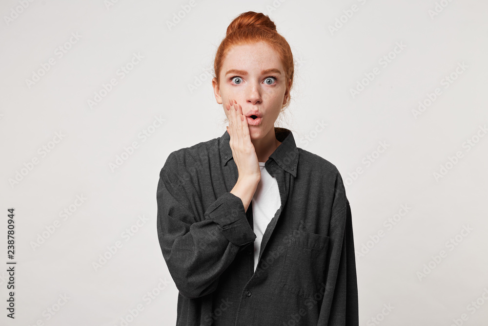 Young pretty woman with red hair, opens mouth with shock, keeps palm on cheek, finds out awful bad news, wears black shirt, being frightened by something, isolated over white background. Omg concept Young pretty woman with red hair, opens mouth with shock, keeps palm on cheek, finds out awful bad news, wears black shirt, being frightened by something, isolated over white background. Omg concept