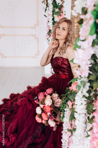 Stunning woman in red burgundi dress stands with colorful bouquet of penies in a room full of flowers photo