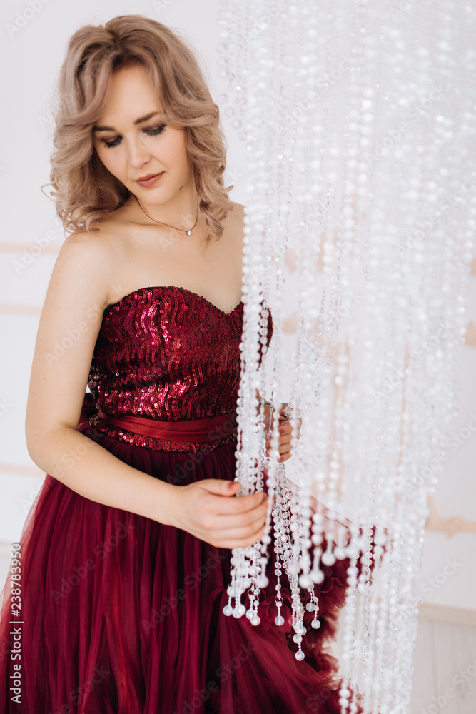 Fototapeta premium Adorable woman in red burgundi dress poses in a bright luxury room with large chandelier