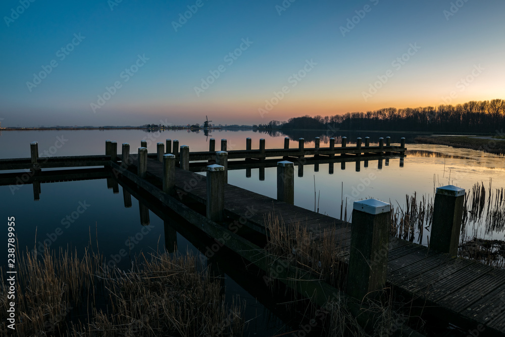 A pier on a lake with a windmill in the distance after sunset. Photograph was taken near lake Rottemeren in The Netherlands.