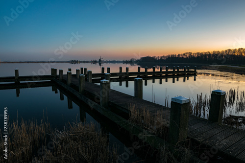 A pier on a lake with a windmill in the distance after sunset. Photograph was taken near lake Rottemeren in The Netherlands.