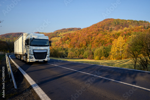 White truck driving on asphalt road around the mountain with deciduous trees in autumn colors