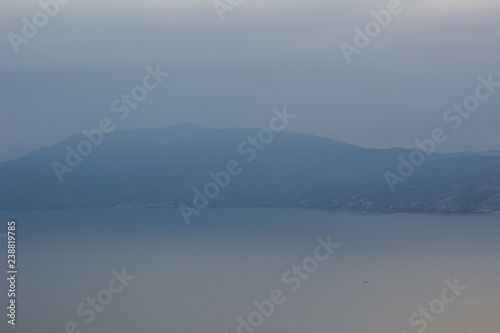 abstract nature landscape wallpaper background pattern of mountain ridge unfocused silhouette on big lake with calm water surface shore line in morning cloudy and foggy weather time