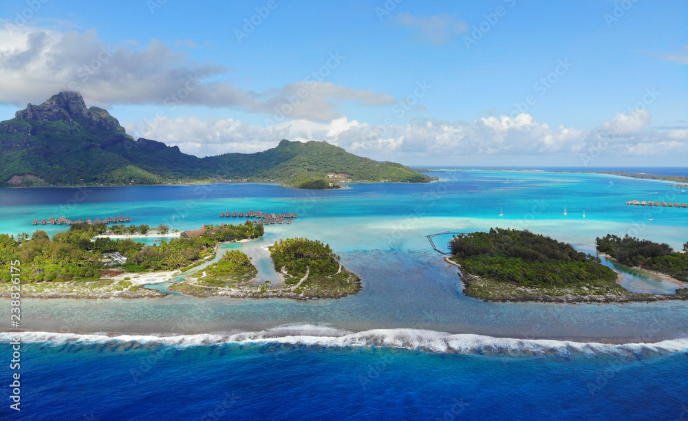 Aerial panoramic landscape view of the island of Bora Bora in French Polynesia with the Mont Otemanu mountain surrounded by a turquoise lagoon, motu atolls, reef barrier, and the South Pacific Ocean