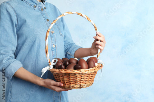 Woman holding wicker basket with chocolate Easter eggs on color background, closeup. Space for text