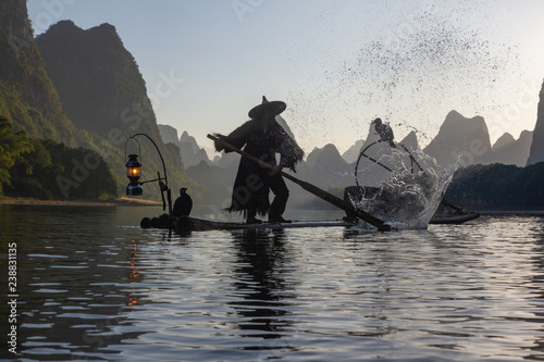 Silhouette of Chinese cormorant fisherman paddling a raft on a lake in Guilin China. Two cormorant birds on raft and lantern lit on front of raft.