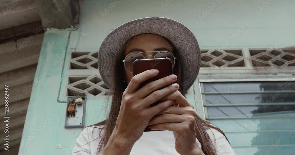 Young woman in stylish hat browsing modern smartphone while standing near weathered building