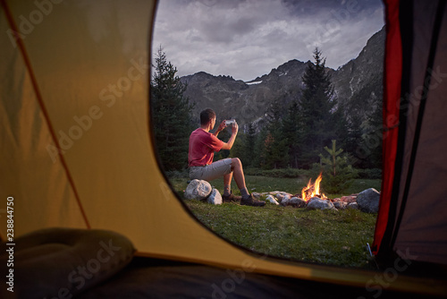 View from inside tent at sunset. Man hiker sitting at bonfire with cellphone