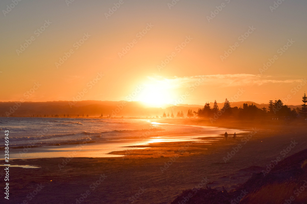 Two people walk the beach as the huge orange sun sets in the background in Gisborne, New Zealand.