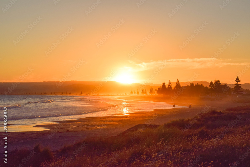 Couple walk along the empty beach at sunset in Gisborne, New Zealand. - obrazy, fototapety, plakaty 