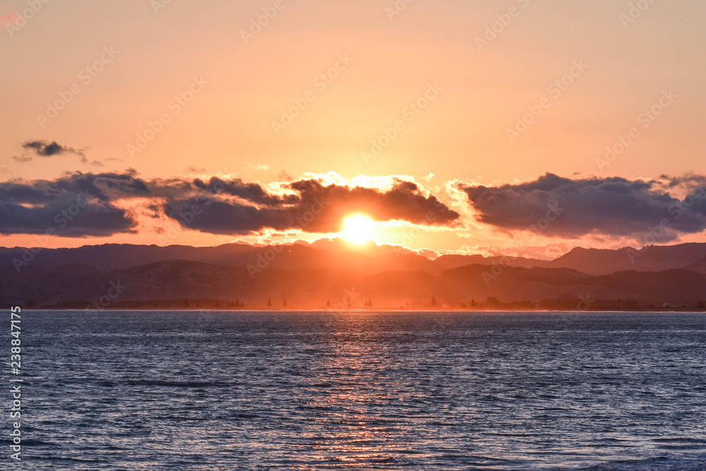 A circuar orange sunset shaped by surrounding clouds at the beach in Gisborne, New Zealand.