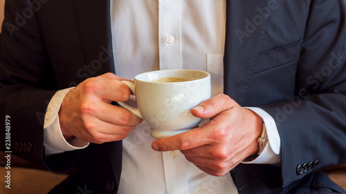 Man holding a cup of coffee with cream in white ceramic cup, dressed in white dress shirt and black business suit and wearing a watch