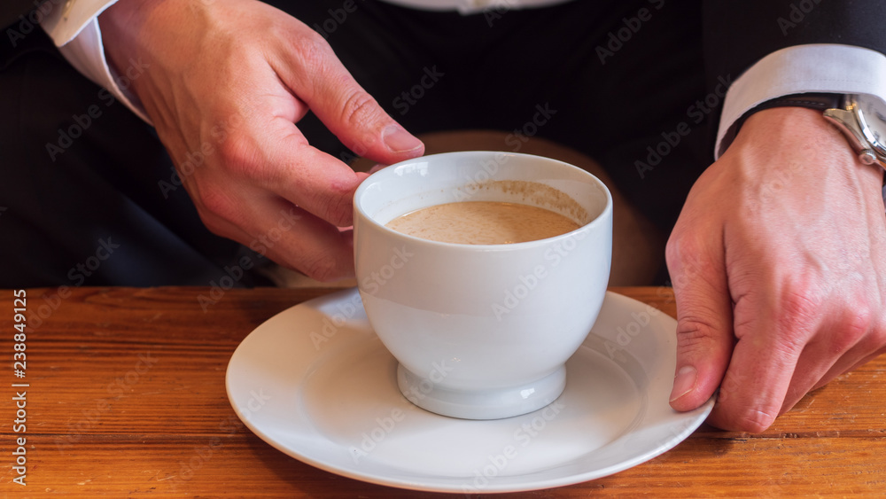 Man sitting at a table drinking coffee with cream in a white ceramic cup on a small white ceramic plate, wearing white dress shirt, black business suit and a watch Man sitting at a table drinking coffee with cream in a white ceramic cup on a small white ceramic plate, wearing white dress shirt, black business suit and a watch