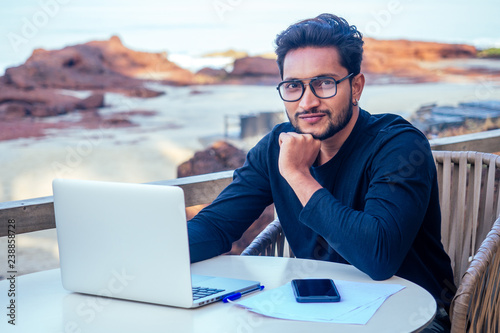 Fotografie, Tablou handsome and successful indian man in a stylish well-dressed freelancer working with a laptop on the beach