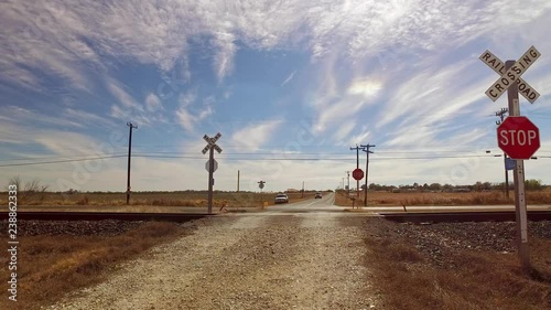 Car approaching a railroad crossing out in the middle of nowhere, long road with wide open fields and blue sunny skies.
