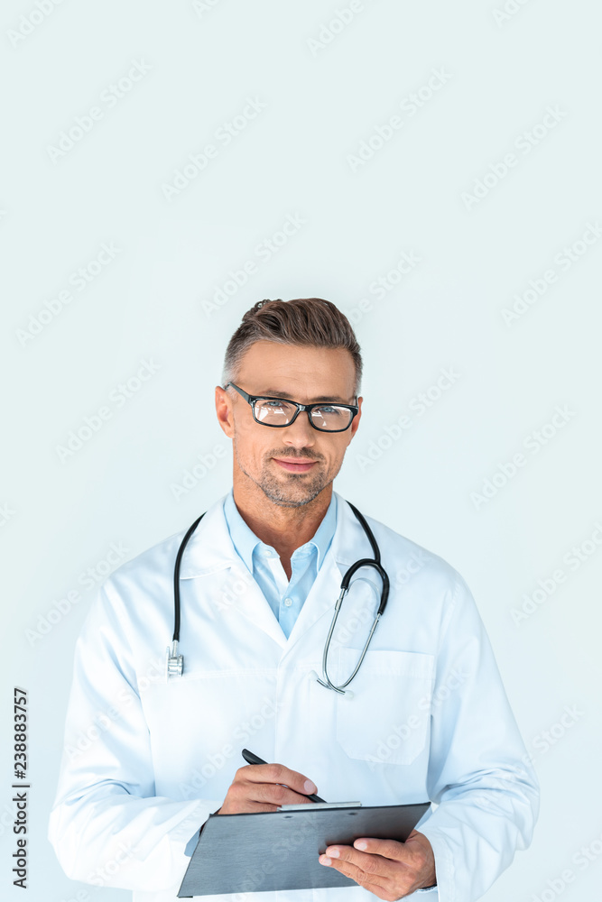 handsome doctor in glasses with stethoscope on shoulders writing something in clipboard and looking at camera isolated on white