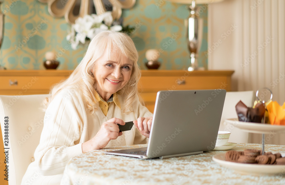 Smiling senior woman uses laptop and credit card while sitting at the table Smiling senior woman uses laptop and credit card while sitting at the table