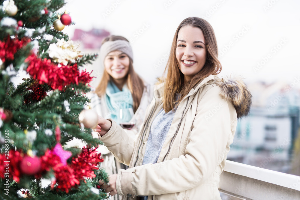 Young girlfriends smiling near Christmas tree on balcony Young girlfriends smiling near Christmas tree on balcony