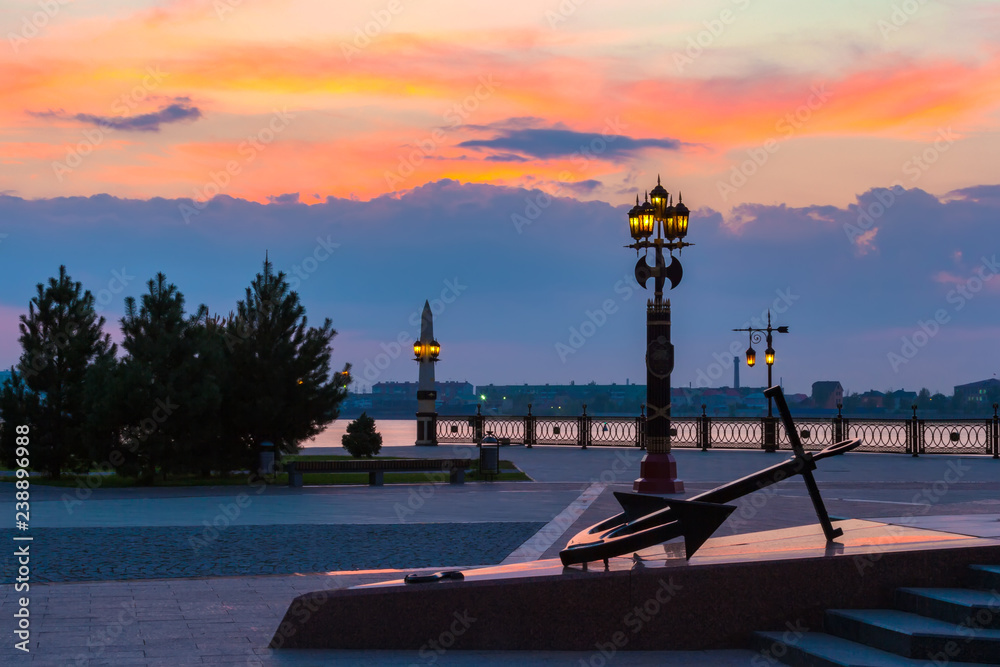 Decorative lamppost with anchor on the square in front of the Volga river bank during sunset with a beautiful colorful sky and an outgoing orange sun. Petrovskaya embankment, Astrakhan, Russia.