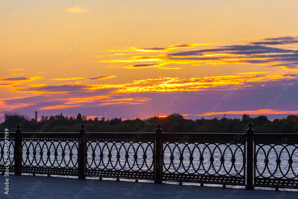 Pedestrian promenade on the Volga River in colorful sunset, silhouette of a fence in dark evening, river at dusk and the sky in bright colors. Petrovskaya embankment, Astrakhan, Russia.