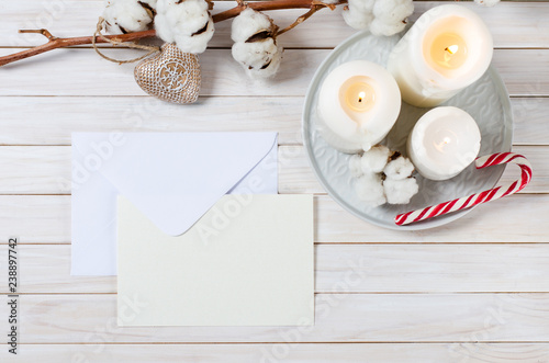 blank white christmas greeting card and envelope with cotton branch, candles and candy cane on white wooden background. mock up. top view with copy space